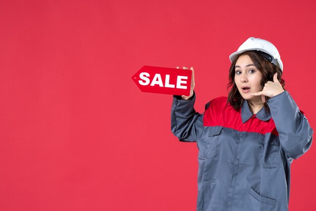 Halbkörperaufnahme einer lächelnden Arbeiterin in Uniform, die einen Schutzhelm trägt und ein Verkaufssymbol zeigt, das mich auf isoliertem rotem Hintergrund anruft