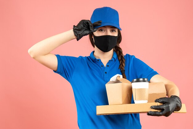 Halbkörperaufnahme eines Kuriermädchens mit medizinischer Maske und Handschuhen, das Bestellungen auf pastellfarbenem Pfirsichhintergrund hält holding