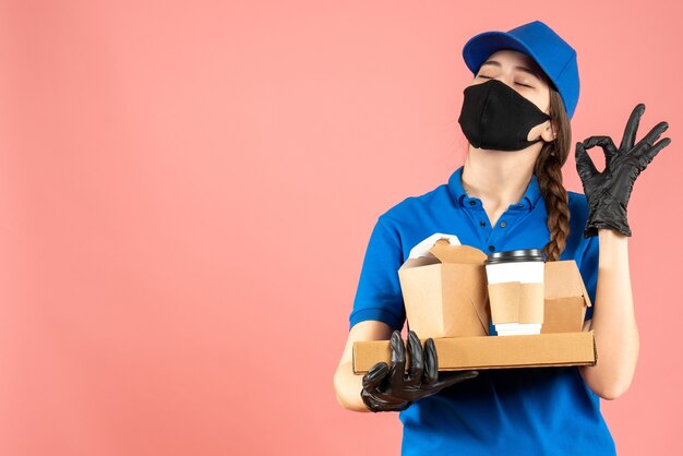 Halbkörperaufnahme eines Kuriermädchens mit medizinischer Maske und Handschuhen, das Bestellungen hält, die eine Brillengeste auf pastellfarbenem Pfirsichhintergrund machen