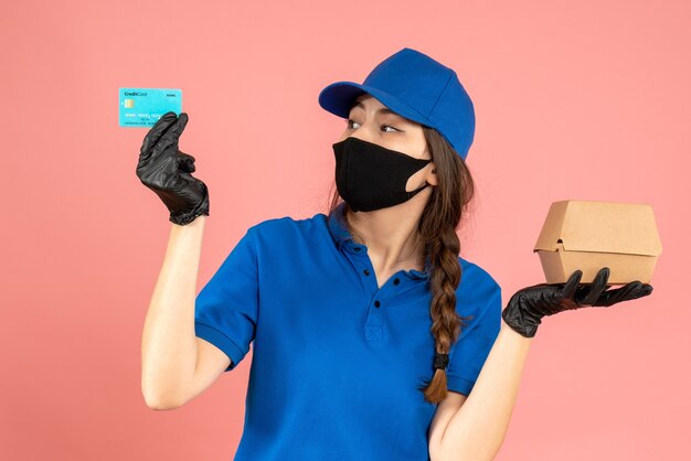 Halbkörperaufnahme eines Kuriermädchens mit schwarzen medizinischen Maskenhandschuhen mit Bankkarte und kleiner Schachtel auf pastellfarbenem Pfirsichhintergrund
