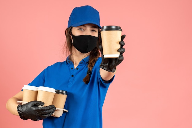 Halbkörperaufnahme eines selbstbewussten Kuriermädchens mit schwarzen medizinischen Maskenhandschuhen, die Kaffee auf pastellfarbenem Pfirsichhintergrund halten holding