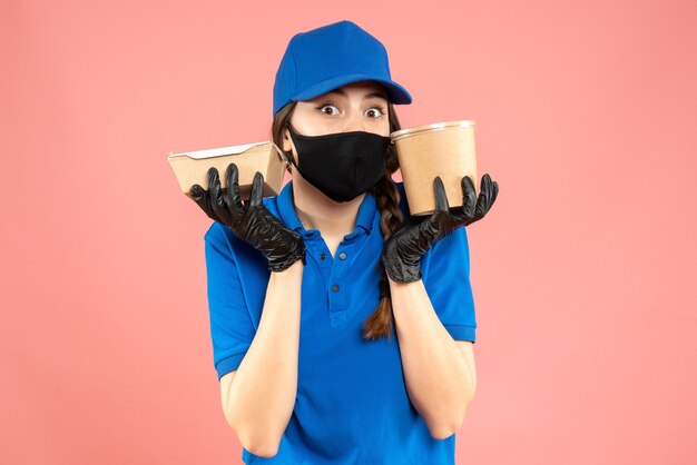 Halbkörperaufnahme eines überraschten Kuriermädchens mit medizinischer Maske und Handschuhen, das eine kleine Kaffeebox auf pastellfarbenem Pfirsichhintergrund hält