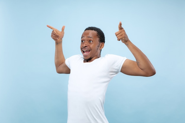 Halblanges Nahaufnahmeporträt des jungen Afroamerikanermannes im weißen Hemd auf blauem Hintergrund. Menschliche Gefühle, Gesichtsausdruck, Anzeige, Konzept. Feiern, verwundert, erstaunt, schockiert, verrückt glücklich.