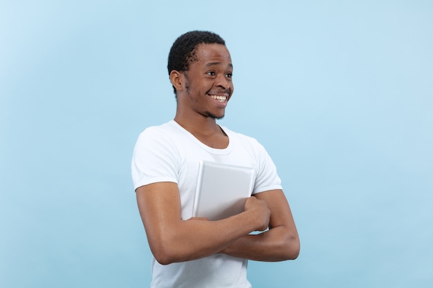 Halblanges Nahaufnahmeporträt des jungen Afroamerikaners im weißen Hemd auf blauem Raum. Eine Tablette halten und lächeln