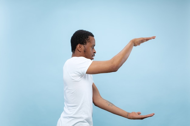 Halblanges Nahaufnahmeporträt des jungen Afroamerikaners im weißen Hemd auf blauer Wand. Menschliche Emotionen, Gesichtsausdruck, Anzeigenkonzept. Halten Sie eine leere Leiste und kopieren Sie den Text für Ihren Text.