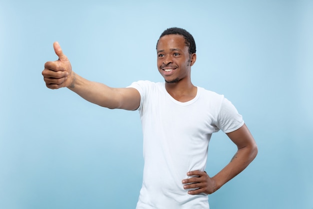 Halblanges Nahaufnahmeporträt des jungen afroamerikanischen männlichen Modells im weißen Hemd auf blauem Raum. Menschliche Emotionen, Gesichtsausdruck, Anzeigenkonzept. Zeigt das Zeichen von OK an