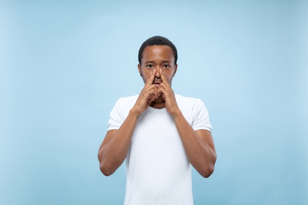 Halblanges Nahaufnahmeporträt des jungen afroamerikanischen männlichen Modells im weißen Hemd auf blauer Wand. Menschliche Emotionen, Gesichtsausdruck, Anzeigenkonzept. Das Gesicht mit den Händen bedeckend, sieht ernst aus.