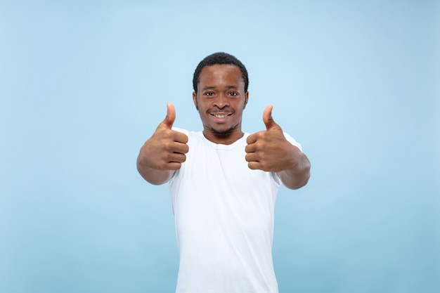 Halblanges Nahaufnahmeporträt des jungen afroamerikanischen männlichen Modells im weißen Hemd auf blauer Wand. Menschliche Emotionen, Gesichtsausdruck, Anzeigenkonzept. Zeigt das Zeichen von OK an. nett.