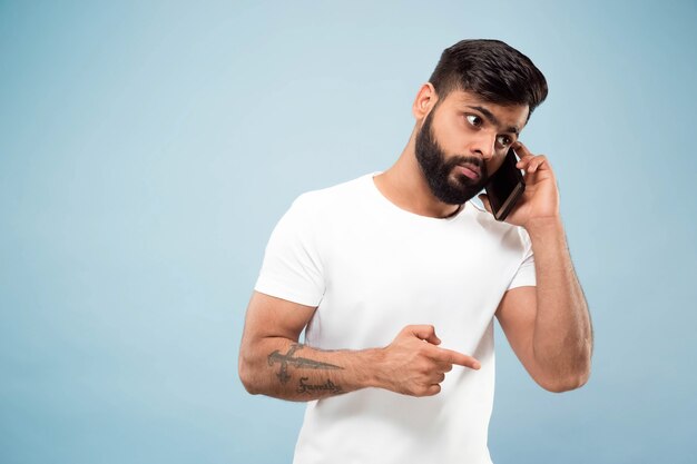 Halblanges Nahaufnahmeporträt des jungen Hindu-Mannes im weißen Hemd auf blauem Hintergrund. Menschliche Emotionen, Gesichtsausdruck, Anzeigenkonzept. Negativer Raum. Auf dem Handy sprechen, Sprachnachrichten aufzeichnen.