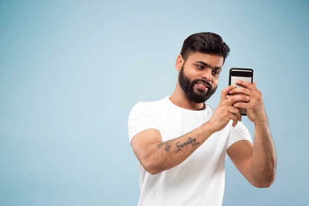 Halblanges Nahaufnahmeporträt des jungen Hindu-Mannes im weißen Hemd auf blauem Hintergrund. Menschliche Emotionen, Gesichtsausdruck, Anzeigenkonzept. Negativer Raum. Selfie oder Videoblog, Vlog, Chating machen.