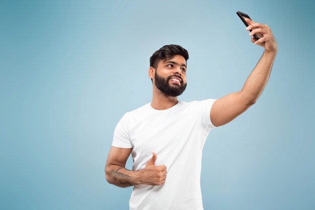 Halblanges Nahaufnahmeporträt des jungen Hindu-Mannes im weißen Hemd auf blauem Hintergrund. Menschliche Emotionen, Gesichtsausdruck, Anzeigenkonzept. Negativer Raum. Selfie oder Videoblog, Vlog, Chating machen.