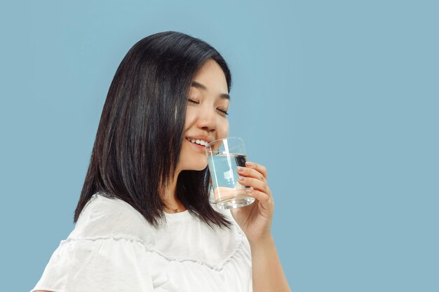 Halblanges Porträt der koreanischen jungen Frau auf blauem Raum. Weibliches Modell im weißen Hemd. Trinkwasser genießen.