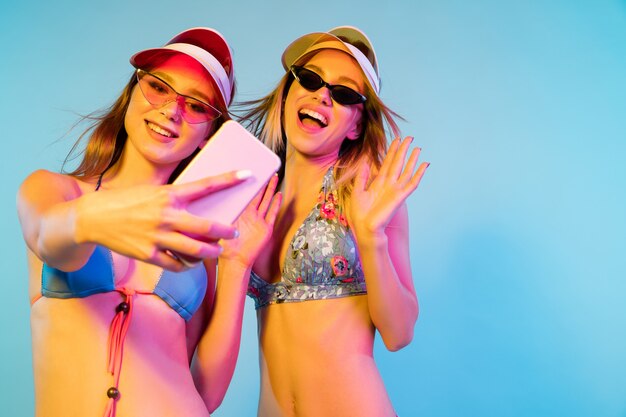 Kostenloses Foto halblanges porträt der schönen jungen mädchen einzeln auf blauem studiohintergrund im neonlicht. frauen posieren im modischen body. gesichtsausdruck, sommer, wochenendkonzept. trendige farben.