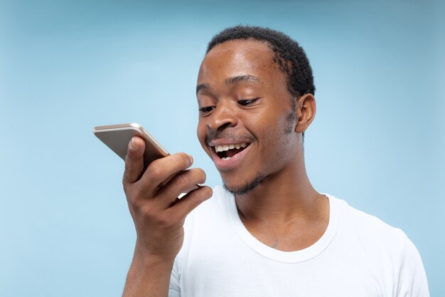 Halblanges Porträt des jungen Afroamerikanermannes im weißen Hemd auf blauem Hintergrund. Menschliche Emotionen, Gesichtsausdruck, Anzeige, Verkaufskonzept. Halten Sie ein Smartphone, sprechen Sie oder zeichnen Sie eine Sprachnachricht auf.