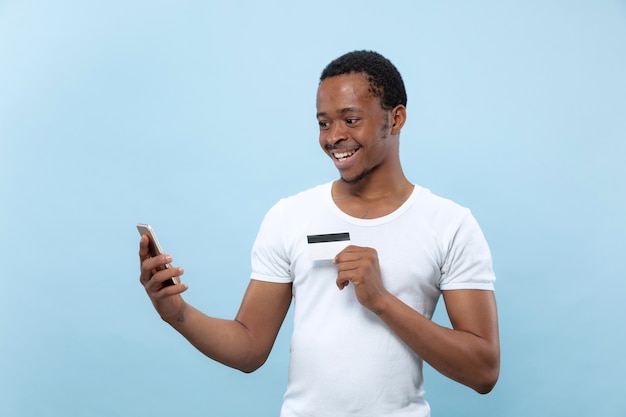 Halblanges Porträt des jungen Afroamerikanermannes im weißen Hemd, das eine Karte und ein Smartphone auf blauem Raum hält