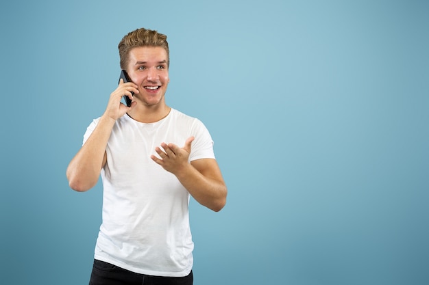 Halblanges Porträt des kaukasischen jungen Mannes auf blauem Studiohintergrund. Schönes männliches Modell im Hemd. Konzept der menschlichen Emotionen, Gesichtsausdruck, Verkauf, Anzeige. Am Telefon sprechen, sieht glücklich aus.