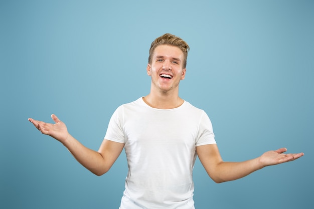 Halblanges Porträt des kaukasischen jungen Mannes auf blauem Studiohintergrund. Schönes männliches Modell im Hemd. Konzept der menschlichen Emotionen, Gesichtsausdruck, Verkauf, Anzeige. Etwas zeigen und zeigen.