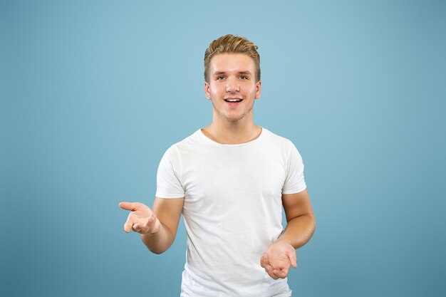 Halblanges Porträt des kaukasischen jungen Mannes auf blauem Studiohintergrund. Schönes männliches Modell im Hemd. Konzept der menschlichen Emotionen, Gesichtsausdruck, Verkauf, Anzeige. Etwas zeigen und zeigen.