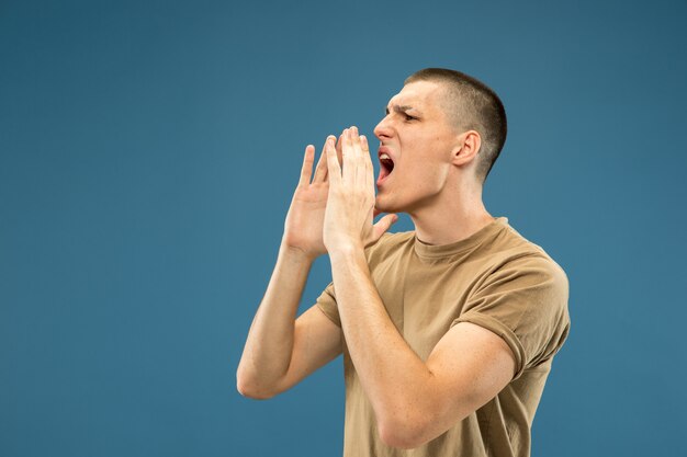 Halblanges Porträt des kaukasischen jungen Mannes auf blauem Studiohintergrund. Schönes männliches Modell im Hemd. Konzept der menschlichen Emotionen, Gesichtsausdruck, Verkauf, Anzeige. Jemanden anrufen, schreien.