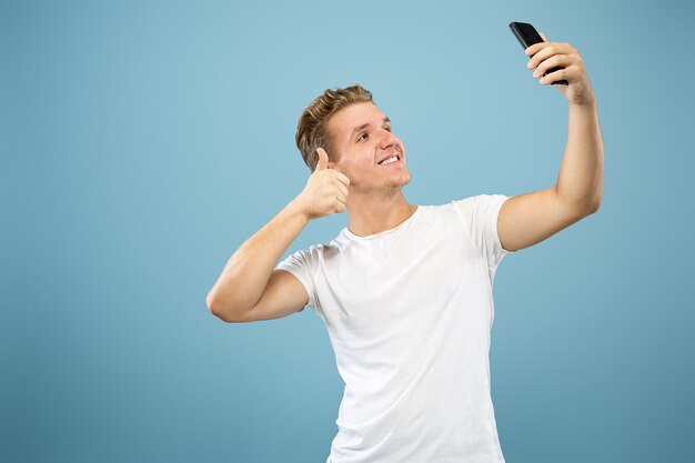 Halblanges Porträt des kaukasischen jungen Mannes auf blauem Studiohintergrund. Schönes männliches Modell im Hemd. Konzept der menschlichen Emotionen, Gesichtsausdruck, Verkauf, Anzeige. Selfie oder Inhalt für vlog erstellen.