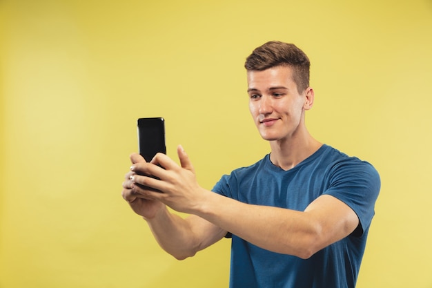 Halblanges Porträt des kaukasischen jungen Mannes auf gelbem Studiohintergrund. Schönes männliches Modell im blauen Hemd. Konzept menschlicher Emotionen, Gesichtsausdruck. Selfie oder Vlog zu machen, sieht glücklich aus.