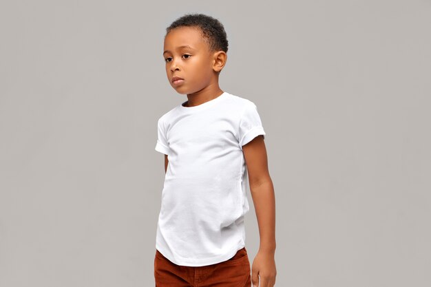 Halbprofilbild des lässig gekleideten afrikanischen Jungen im weißen T-Shirt mit ruhigem selbstbewusstem Gesichtsausdruck, der isoliert gegen leere Wand mit Kopienraum für Ihre Informationen aufwirft