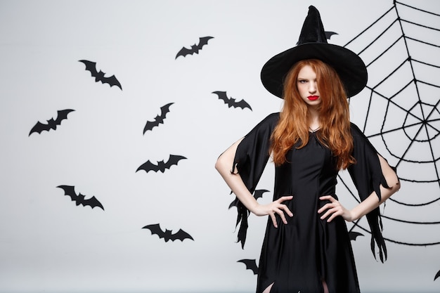 Halloween-Hexe-Konzept Halloween-Hexe, die mit ernstem Ausdruck über dunkelgrauer Wand mit Fledermaus und Spinnennetz posiert