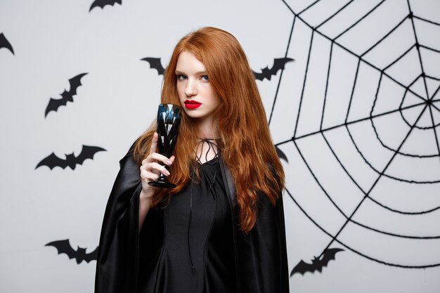 Halloween-Hexe-Konzept - Happy Halloween-Hexe, die ein Glas blutigen Rotweins über dunkelgrauer Wand mit Fledermaus und Spinnennetz hält.