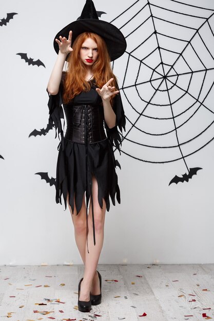 Halloween-Hexe-Konzept in voller Länge Halloween-Hexe, die Zaubersprüche mit ernstem Ausdruck über dunkelgrauer Wand mit Fledermaus und Spinnennetz wirft
