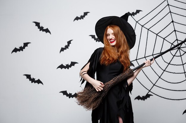 Halloween-Konzept glückliche elegante Hexe genießt das Spielen mit Besenstiel-Halloween-Party über grauer Wand