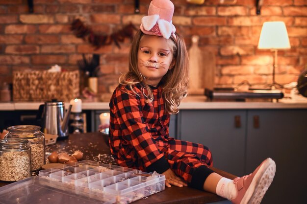 Halloween-Konzept. Süßes kleines Mädchen mit Kaninchen-Make-up-Hut und Schlafanzug, das morgens auf dem Tisch mit verstreuten Speisen in der Küche im Loft-Stil sitzt.