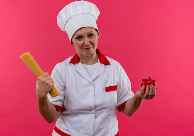 Halten der Köchin mittleren Alters in der Kochuniform, die Spaghetti und Pfeffer auf isolierter rosa Wand hält