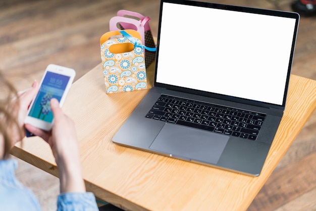 Hand der Frau, die Smartphone mit Laptop des leeren Bildschirms auf Holztisch hält