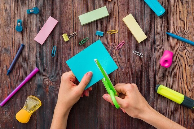 Hand der Person, die blaues Papier über dem Schulzubehör auf Holztisch schneidet