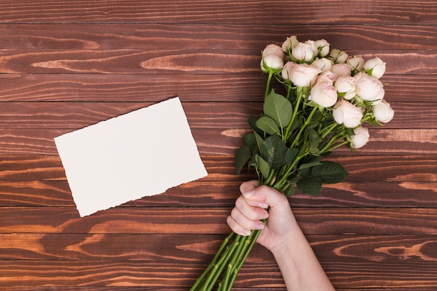 Hand der Person, die Bündel weiße Rosen hält; leeres Papier über Schreibtisch aus Holz