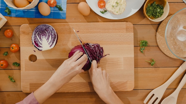 Hand des jungen asiatischen Frauenkochs halten Messer, das Rotkohl auf Holzbrett schneidet