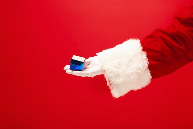 Hand des Weihnachtsmannes, der ein Geschenk auf rotem Hintergrund hält. Jahreszeit, Winter, Feiertag, Feier, Geschenkkonzept