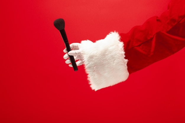 Hand des Weihnachtsmannes, der einen Make-up-Pinsel für Puder auf rotem Hintergrund hält. Jahreszeit, Winter, Feiertag, Feier, Geschenkkonzept