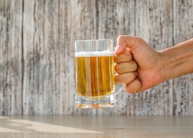 Hand, die Bier in einem Glasbecher auf grungy und hellem Tisch, Seitenansicht hält.