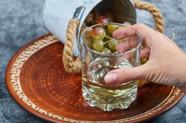 Hand, die ein Glas Weißwein und einen kleinen Eimer Trauben auf einem Marmorhintergrund hält. Hochwertiges Foto