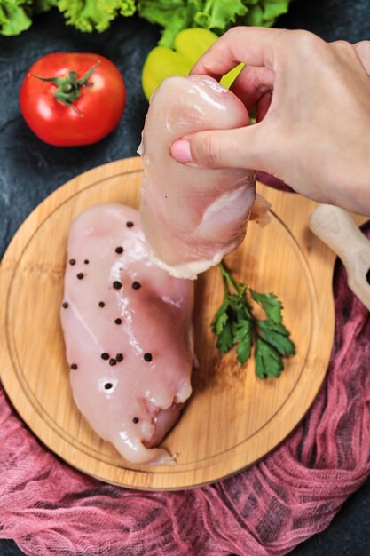 Hand, die ein Stück rohes Hühnerfilet von der Holzplatte mit frischem Gemüse hält.