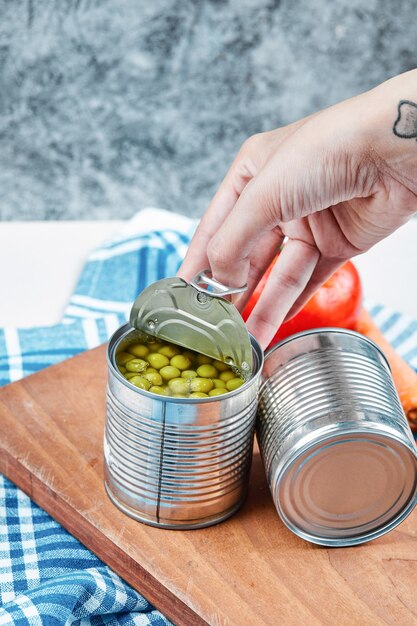 Hand, die eine Dose gekochte grüne Erbsen auf einem weißen Tisch mit Gemüse und Tischdecke hält.