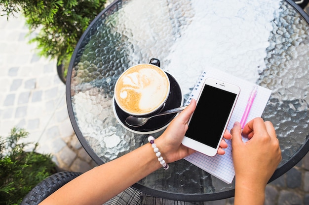 Hand, die Mobiltelefon mit Kaffeetasse auf Glastisch hält