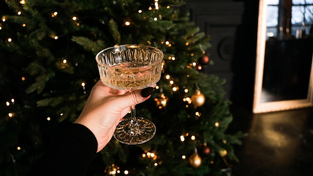 Hand, die stilvolles vintage-weinglas auf dem hintergrund der schönen weihnachtsbaumbeleuchtung hält. frohe weihnachten und ein glückliches neues jahr-konzept. schöne ferien