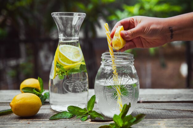 Hand, die Zitrone in ein Glas mit Wasserseitenansicht auf Holz- und Gartentisch drückt