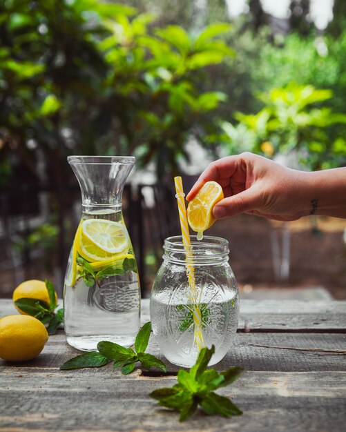 Hand, die Zitrone in Wasser in der Glasglas-Seitenansicht auf Holz- und Gartentisch drückt