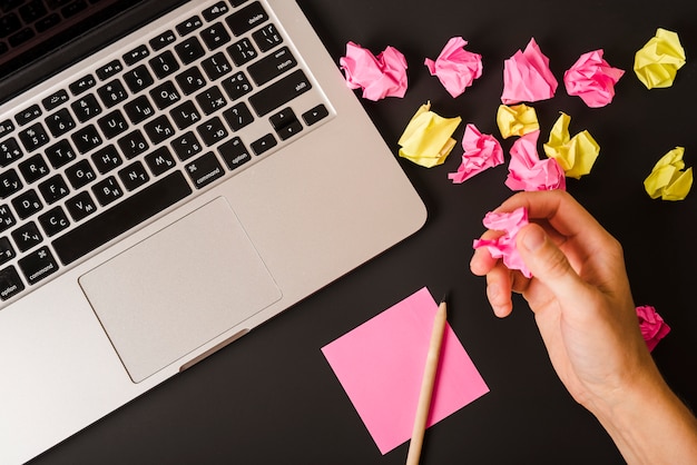 Hand einer Person, die zerknittertes Papier mit Laptop hält; Stift- und Klebepapier auf schwarzem Hintergrund