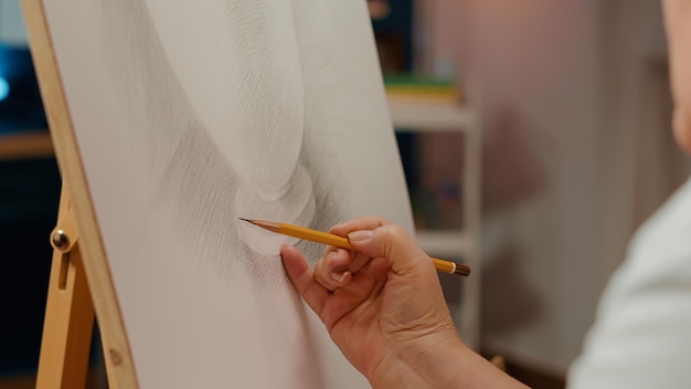 Hand eines professionellen Künstlers, der Vasenmodelldesign mit Bleistift auf weißer Leinwand und hölzerner Staffelei zeichnet. Verwenden Sie kreative Fähigkeiten und künstlerische Werkzeuge, um eine Umrissskizze mit Inspiration zu erstellen. Nahansicht.