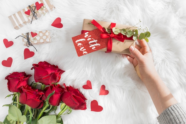 Hand mit Geschenk mit Tag nahe Verzierungsherzen und frischen Blumen auf woolen Bettdecke