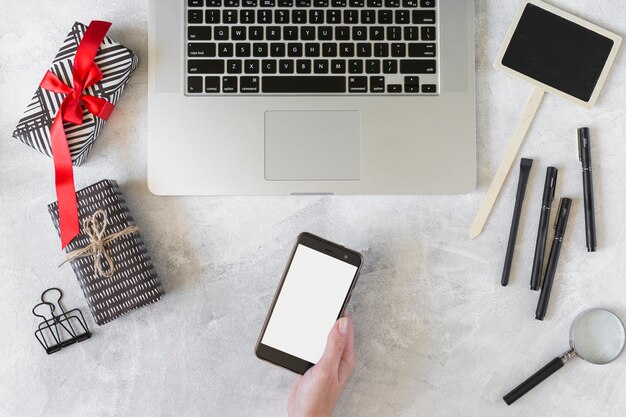 Hand mit Smartphone nahe Laptop, Präsentkartons und Briefpapier